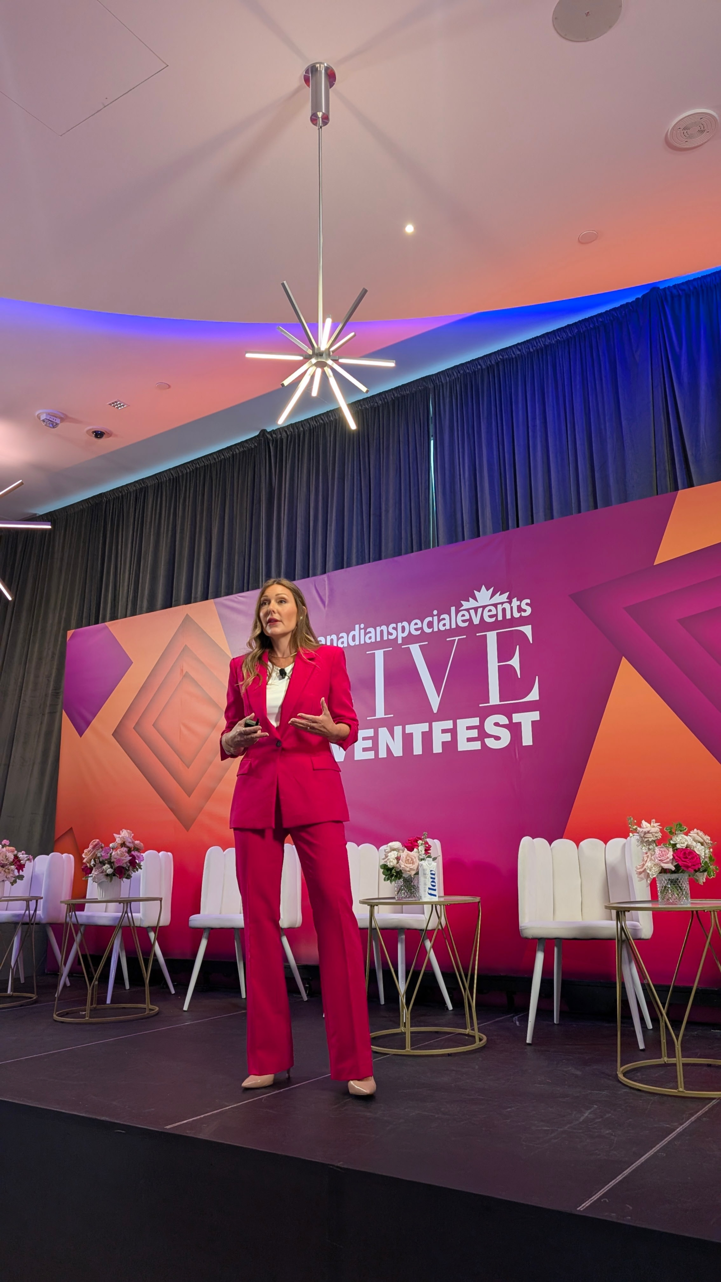 Woman in a pink suit giving a talk on a stage with a vibrant banner behind her that reads 'LIVE' and event seating nearby.